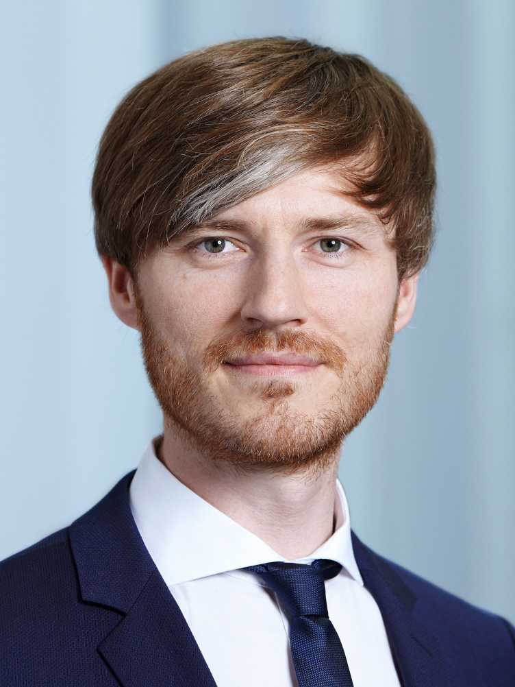 Portrait of Prof. Alexander Mathys with short light brown hair, wearing a navy blue suit, white shirt, and dark tie, smiling slightly at the camera.